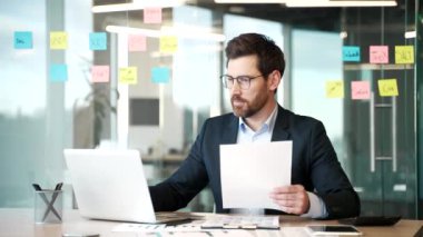 İş yerindeki mali sonuçlardan memnun olan mutlu bir finansör. Gülümseyen işadamı belgelere ve bilgisayara bakıyor ve başarılı yatırımı kutluyor