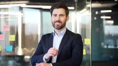 Kendine güvenen işadamı modern ofiste kollarını kavuşturmuş duruyor. Takım elbise giymiş, kameraya gülümsüyor, profesyonellik ve liderlik yayıyor. Kapalı bir şirket ortamı, başarılı bir girişimci