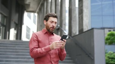 Sakallı erkek girişimci dışarıda telefonu tutarken heyecanını dile getiriyor. Şehir ortamında başarılı haberleri kutlayan kırmızı gömlekli iş adamı, mutluluk ve profesyonel başarı sergiliyor