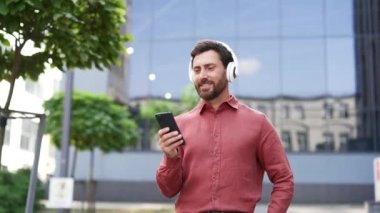 Şehirde kulaklık takmış ve telefon kullanan kendine güvenen bir iş adamı. Gülümserken müziği sever, modern yaşam tarzı dengesini somutlaştırır. Kırmızı gömlek, kentsel geçmiş, profesyonel canlılık.