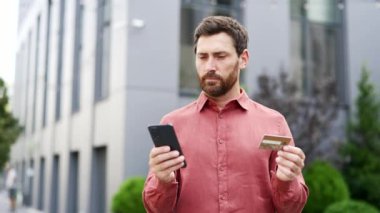 Telefon kullanırken gülümseyen ve şehir merkezinde kredi kartı tutan yakışıklı bir iş adamı. Mobil bankacılık kavramı, online alışveriş, modern teknoloji. Sakallı adam satın almaktan memnun.