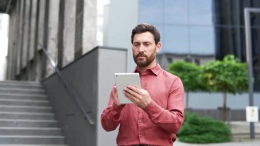 Ofis binasının önünde dijital tablet kullanan kırmızı gömlekli sakallı adam. Yakışıklı erkek mesajlaşması, internette sohbet etmesi, bakması, mesaj yazması. İşletme profesyoneli iş başında.