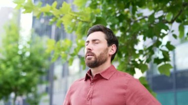 Sakallı bir iş adamı ofis binasının önünde durmuş, temiz havanın tadını çıkarıyor. Kırmızı bir gömlek giyiyor, sakin ve rahat görünüyor. Mutlu, mutlu, gözleri kapalı adam derin derin nefes alıyor. Şehir merkezinde ayakta duruyor.