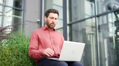 İş adamı şehir ofisinin dışında oturuyor, dizüstü bilgisayarla çalışırken hayal kırıklığı ve hayal kırıklığı gösteriyor. Kırmızı gömlekli sakallı adam bilgisayardan kötü haberler okudu. Erkek rahatsız edici bir haberle sarsıldı