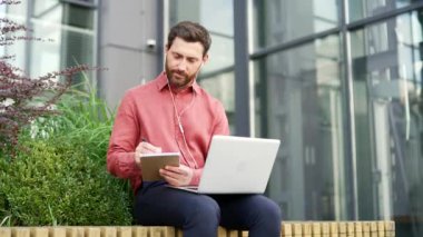 İşadamı dışarıda bankta dizüstü bilgisayarla oturmuş not alıyordu. Kulaklıklı bir adam çevrimiçi video konferansı izliyor ve bilgisayar ekranına bakıyor. Erkek dinleme uzaktan iş eğitimi veya kursu