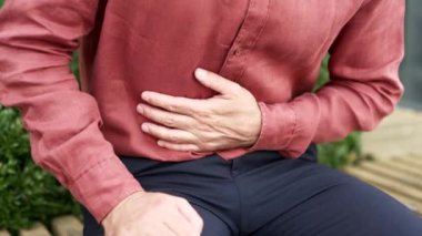 Kapatın. Erkek el, karın masajı yapıyor. Kırmızı gömlekli adam, işyeri binasının yakınındaki bir bankta otururken mide ağrısını hisseder. Hasta adam mide ekşimesi, mide iltihabı ve kabızlıktan muzdarip.