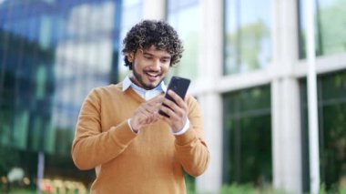 Modern ofis binasının dışındaki akıllı telefonla başarıyı kutlayan mutlu iş adamı. Gündelik giyinen Latin erkek heyecanlı ve neşeli hissediyor. iş başarısı, kentsel yaşam tarzı, olumlu duygular
