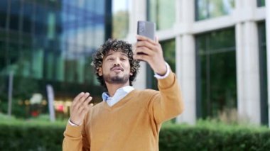 Gülümseyen genç adam modern ofis binasının önünde video görüşmesi yapıyor, el sallıyor. Akıllı telefondan konuşma, dostane tavır. iletişim, teknoloji ve profesyonel etkileşim