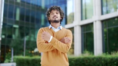 Kendine güvenen genç girişimci ofis çalışanı şehir ortamında gülümsüyor, kollarını kavuşturmuş, sıradan süveter giymiş, arka plandaki modern binadaki kameraya bakan erkek işadamı.