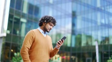 Modern iş yeri binasının yakınında gündelik yürüyüşler yapan yakışıklı iş adamı akıllı telefonu elinde tutuyor. Genç yetişkin girişimci şehir dışında bir cep telefonu kullanıyor.