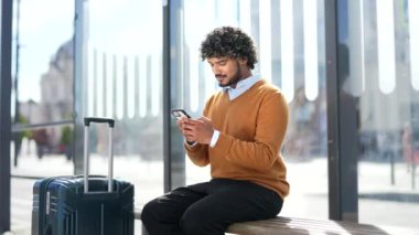 Otobüs durağında akıllı telefon kullanarak bankta oturan bir gezgin. Bavullu gezgin teknoloji ve iletişimden zevk alıyor. Telefon ekranına bakıyor. Genç adam nakil için bekliyor.