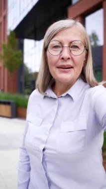 POV gülümseyen, gri saçlı olgun bir kadın. Dışarıdaki kameraya bakmak için telefon kullanıyor. Yaşlı bir kadın el sallıyor, şehir sokağında sevdikleriyle mutluluk ve bağ kuruyor. Dikey