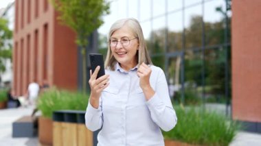 Olgun bir kadın şehir binasının önünde dikilirken telefonla video görüşmesi yapıyor. Yaşlı mutlu kadın sevdikleriyle çevrimiçi konuşuyor. Dalgalar mutlu, modern teknoloji ve iletişim sergiliyor.