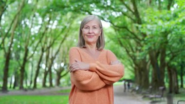 Huzurlu park ortamında kendine güvenen yaşlı bir kadın kameraya bakıyor, mutluluk ve memnuniyet gösteriyor. Turuncu kazaklı olgun bir kadın çapraz kollarıyla doğanın tadını çıkarıyor.