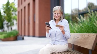 Dışarıda bankta oturan yaşlı bir kadın telefon kullanıyor ve kredi kartı tutuyor. Gri saçlı olgun bir kadın çevrimiçi alışveriş, modern yaşam tarzı ve açık hava kentsel ortamında dijital kolaylıktan hoşlanıyor.
