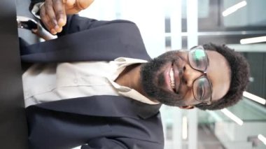 POV dikey. Resmi takım elbiseli, gülümseyen Afro-Amerikan bir işadamı iş yerinde otururken telefonla konuşuyor. Siyah adam kameraya bakarak iletişim kuruyor. Çevrim içi sohbet