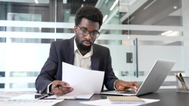 Düşünceli afro-amerikan iş adamı bir iş yerinde masa başında otururken bilgisayardaki verileri kontrol ederek finansal belgeleri analiz ediyor. Finansör mali raporla ilgilenir
