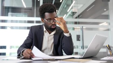 Çok çalışan işadamı, iş yerindeki masasında otururken evrak işi yapmaktan yoruldu. Resmi takım elbiseli bitkin siyah işçi elinde hayal kırıklığına uğramış gibi görünen belgeler tutuyor.