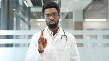 Beyaz önlüklü Afro-Amerikalı erkek doktor hastanedeki klinikte parmağını kaldırarak hayır diyor. Tıp doktoru bir yasaklama, inkar, anlaşmazlık jesti gösteriyor.