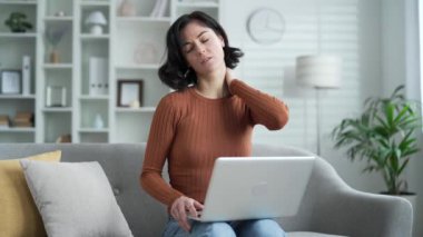 Evde oturma odasındaki kanepede otururken dizüstü bilgisayarla çalışırken boynu ağrıyan genç bir kadın. Üzgün kadın masajları ve ağrıyan kasları elleriyle ovuyor, esnetiyor.