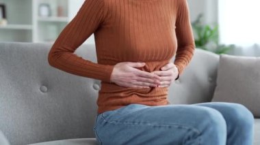 Kapatın. Karın masajı yapan kadın elleri. Oturma odasındaki kanepede otururken karın ağrısı çeken genç bir kadın. Zehirlenme, kabızlık, spazm ya da regl olmuş.