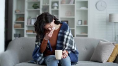 Evdeki oturma odasında elinde kupa tutarken battaniyeyle hapşıran genç bir kadın. Hasta kadın soğuk, burnu akan ve grip olmuş. Yorgun kadın burnunu mendille siler.