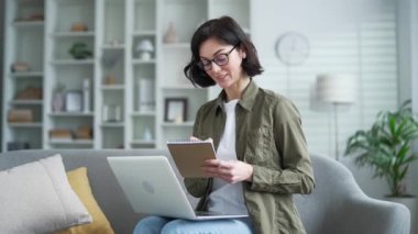 Genç bir kadın rahat koltukta dizüstü bilgisayar ve defter kullanarak evde uzaktan öğrenmeyi seviyor. Çevrimiçi çalışmalara odaklanmış, modern apartmandaki işe odaklanmış, verimli bir atmosfer yaratıyor.