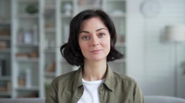 Modern, rahat bir evde otururken kısa siyah saçlı hafifçe gülümseyen genç bir kadının portresi. Oturma odasında kameraya bakarak poz veren kendinden emin mutlu bir kadının vesikalığı. Kapat.