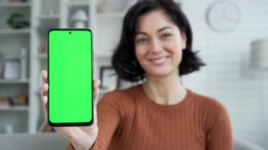Evdeki oturma odasındaki kanepede oturan yeşil ekranlı cep telefonunu gösteren mutlu genç kadın. Renk anahtarı, reklam için dikey şablon düzeni. Gülümseyen kadın kamerası akıllı telefon tutuyor.