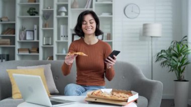 Ev ortamında telefonla pizza yiyen genç bir kadın. Rahat bir koltukta pizza kutusuyla oturmak, gülümseyen bir kadın hızlı yemek teslimatından mutlu olur. Eve yemek götürmek için.