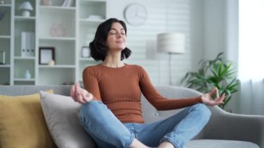 Sakin genç bayan meditasyon evdeki oturma odasındaki kanepede bacak bacak üstüne atarak oturuyor. Mutlu güzel kadın yoga yapıyor, derin derin nefes alıyor, rahatlıyor. Kapalı gözler, sükunet, huzur, sükunet hissi yaratıyor.