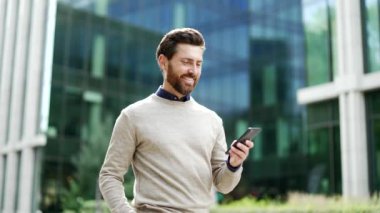 İş adamı dışarıda akıllı telefon tutarak yürüyor, internette gezinirken ya da sohbet ederken gülümsüyor. Şehir yaşamından zevk alan bir erkek profesyonel iş yeri yakınlarındaki caddede mesajları kontrol ediyor.