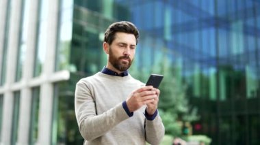 Mutlu iş adamı bir iş yeri binasının yakınındaki caddede duran telefonu kullanıyor. Gülümseyen, sakallı, mesajlaşan, internette sohbet eden, internette gezinen, akıllı telefondan mesajlar yazan bir erkek.