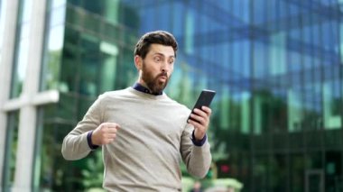 Neşeli iş adamı telefonda harika haberler okuduktan sonra modern ofis binasının dışındaki başarısını kutluyor. Zaferi ve heyecanı ifade etmek. Başarı kavramı, iş zaferi ve olumlu duygu