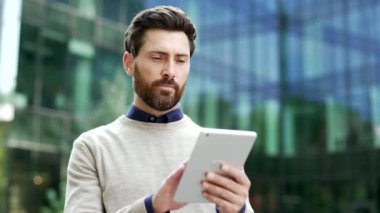 Kendine güvenen ciddi işadamı modern şehir ofis binasının dışında dijital tablet kullanıyor. Şehir sokaklarında teknolojiye odaklanmış durumda. Düşünceli bir adam internette mesajlaşıyor, sohbet ediyor, internette geziniyor.