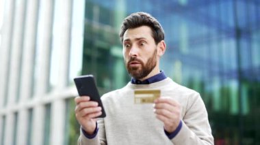 Elinde telefon ve kredi kartı olan işadamı dolandırıcılıktan endişeli görünüyor. Şehirde durmuş endişe ve belirsizliği dile getiriyorsun. Erkek, kafa karışıklığı yaşıyor. Çevrimiçi aldatmanın muhtemel kurbanı.