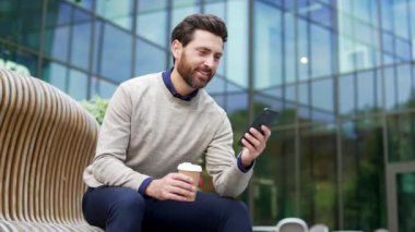 Profesyonel bir erkek, elinde kahveyle bankta oturuyor ve şehir ortamında telefon kullanıyor. Günlük kıyafetlerle gezen iş adamı ara veriyor, mesajları kontrol ediyor, ofis binalarıyla çevrili.