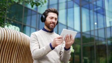 Modern binanın dışındaki bankta otururken dijital tablette video izlemekten zevk alan mutlu erkek profesyonel. Yüzünde kulaklıkla gülen bir işadamı açık havada cihaz kullanıyor.