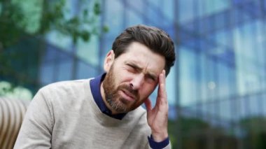 İş yerinin yakınındaki bir bankta oturan baş ağrısından muzdarip iş adamı. Üzgün işçi elleriyle şakaklarına masaj yapıyor. Çok yorgun ve yüksek tansiyonu var. Kapat.