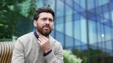 İş yerinin yakınındaki bir bankta otururken boğazı ağrıyan hasta işadamı. Üzgün adam boğazına acı dolu bir ifadeyle dokunuyor, soğuk algınlığı var, virüs ya da grip. Kapat.