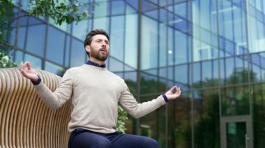 İş adamı, modern cam binanın dışındaki bankta oturmuş meditasyon yapıyor. Olgun erkek profesyonel şehir manzarasında rahatlama ve farkındalık arıyor. Stres atma kavramı, iç huzur.