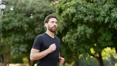 Şehir parkında koşan yakışıklı bir erkek koşucu ve akıllı bir saat. Sakallı sporcu, spor takip cihazı kullanıyor. Sporcu sonuçları kontrol ediyor. Tişörtlü yetişkin adam doğada koşmaktan hoşlanıyor.