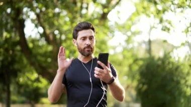 Şehir parkında telefon görüşmesi yapan aktif bir erkek. Egzersiz yaparken video aracılığıyla iletişim kurmak için akıllı telefon kullanan siyah spor kıyafetli biri. Adam yürürken konuşuyor.
