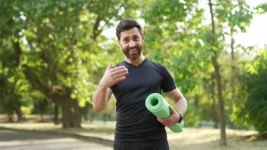 Sporcu adam güvenle gülümseyerek kentteki parkta kameraya bakarak yoga minderini tutarken kendisine katılmak istiyor. Sporcu açık hava fitness aktivitelerini, eğitimi ve aktif bir yaşam tarzını sürdürmeyi teşvik ediyor