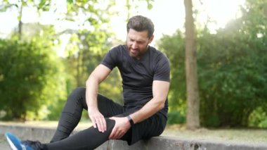 Yetişkin sakallı erkek sporcu, şehir parkında otururken kas ağrısı ve rahatsızlıktan muzdarip. Adam dışarıda idmandan sonra diz ağrısı çekiyor. Travma yaralanmasını hafifletmek için masaj yapıyor ve esniyor.