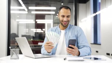 Mutlu Afro-Amerikan iş adamı elinde nakit para banknotları ve iş yerindeki kameralara bakan bir hayran tutuyor. Erkek, akıllı telefonunda bir uygulama kullanarak para kazanmaktan memnun.