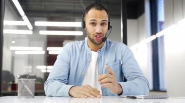 Webcam görüntüsü. Müşteri hizmetlerinde çalışan Afro-Amerikan şirket operatörü kameraya bakan bir video görüşmesinde konuşuyor. Kablosuz kulaklıktaki çağrı merkezi ajanı ofiste çalışıyor. Çevrimiçi danışma