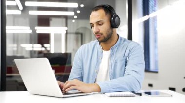 Odaklanmış Afrikalı Amerikalı bilişim uzmanı kablosuz kulaklıklar bir iş yerindeki bir dizüstü bilgisayarla çalışıyor. Ciddi kodlayıcı geliştirici programlıyor, bilgisayardaki bir projeyle meşgul