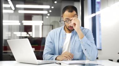 Aşırı çalışan Afrikalı Amerikalı iş adamı göz yorgunluğu ve baş ağrısı çekiyor iş yerindeki bir laptopta çalışıyor. Yorgun siyah adam gözlüğünü ve masaj köprüsünü çıkarıyor.