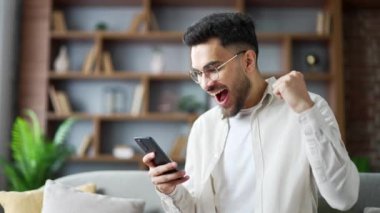 Mutlu yakışıklı adam evdeki oturma odasındaki koltukta otururken cep telefonundan harika haberler aldı. Gülümseyen erkek akıllı telefonda hoş bir mesaj okur, kameraya bakarak başarıyı kutlar. Kapat.
